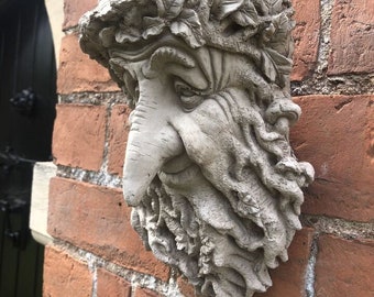 Op de afbeelding: Een verweerde, grijze stenen muursculptuur van een bebaard gezicht met een bladerkroon. De sculptuur is bevestigd aan een rode bakstenen muur. Het gezicht heeft een prominente neus en gedetailleerde gelaatstrekken.