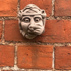 May include: A grey stone sculpture of a face with a crown, set against a red brick wall.