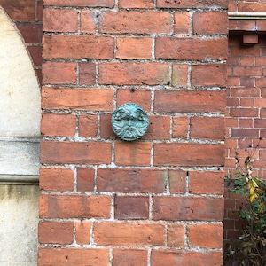 K&ouml;nnte beinhalten: Eine gr&uuml;ne Keramikskulptur eines Gesichts mit einem Blattwerk im Hintergrund, montiert an einer Backsteinmauer.