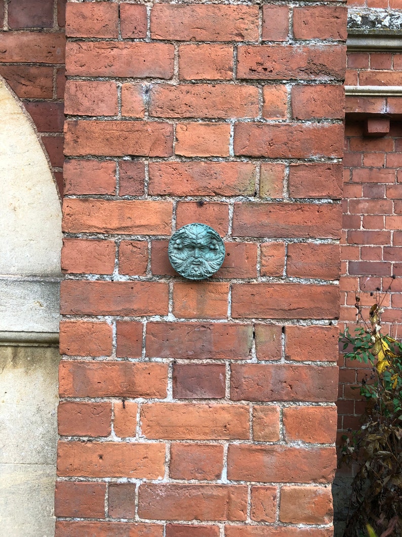 K&ouml;nnte beinhalten: Eine gr&uuml;ne Keramikskulptur eines Gesichts mit einem Blattwerk im Hintergrund, montiert an einer Backsteinmauer.