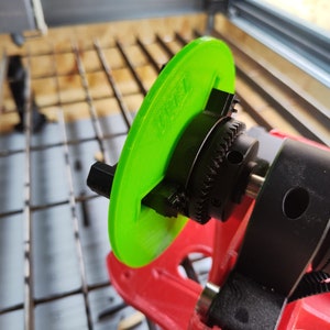 May include: A close-up of a green plastic disc attached to a black metal gear. The disc has a small black plastic piece on the side. The gear is attached to a black metal shaft. The shaft is connected to a black metal base. The base has a black rubber belt wrapped around it.