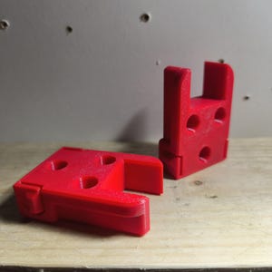 May include: Two bright red, 3D-printed plastic components. One is a rectangular base with a raised section and the other is a matching piece that stands upright. Both have circular holes. The items are on a wooden surface.
