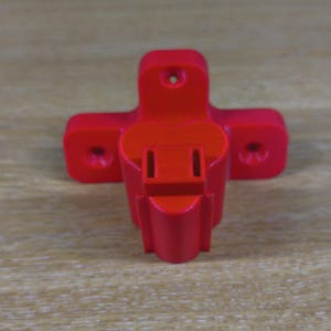 May include: A bright red electrical adapter with a unique cross-shaped design. It features a female plug receptacle and two mounting holes on each side. The adapter is made of a solid, smooth material.