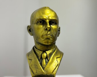 May include: A gold-colored bust sculpture of a man wearing a suit and tie. The sculpture is on a square base.