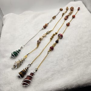 May include: An assortment of handmade jewelry pendants with chains. The pendants feature various gemstones, including green malachite, tiger's eye, and other stones wrapped in silver wire. The chains are gold and silver, with decorative beads.