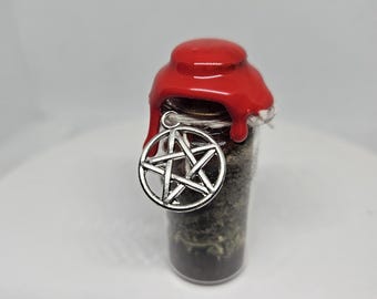 Op de afbeelding: Een klein glazen flesje met een rode waszegel en een zilveren pentagram-hanger. Het flesje is gevuld met gedroogde kruiden.