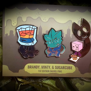 Puede incluir: Tres pines de esmalte sobre una tarjeta con el texto BRANDY, MINTY, & SUGARCUBE 1ST EDITION ENAMEL PINS. Los pines muestran un vaso con l&iacute;quido &aacute;mbar, un personaje con forma de hoja color verde azulado y una criatura marr&oacute;n oscuro.