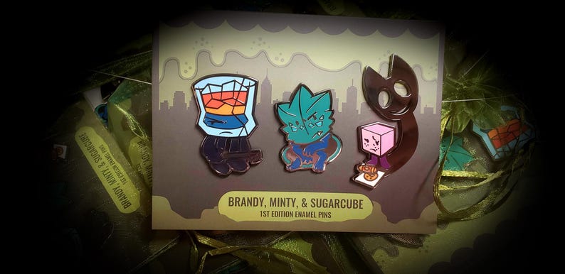 Puede incluir: Tres pines de esmalte sobre una tarjeta con el texto BRANDY, MINTY, & SUGARCUBE 1ST EDITION ENAMEL PINS. Los pines muestran un vaso con l&iacute;quido &aacute;mbar, un personaje con forma de hoja color verde azulado y una criatura marr&oacute;n oscuro.