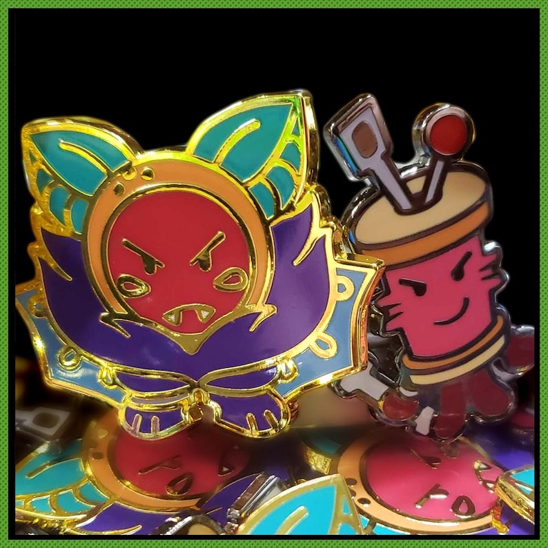 May include: Two enamel pins with gold-toned metal outlines. One pin features a red, purple, and teal creature with pointed ears and a stern expression. The second pin is a pink, cylindrical character with a tan top, holding a brown tool and a red sphere.