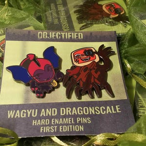 May include: Hard enamel pins featuring two cartoon characters, one with blue wings and the other with a red and brown design. The pins are on a card that reads "WAGYU AND DRAGONSCALE HARD ENAMEL PINS FIRST EDITION". The pins are packaged in green mesh bags.