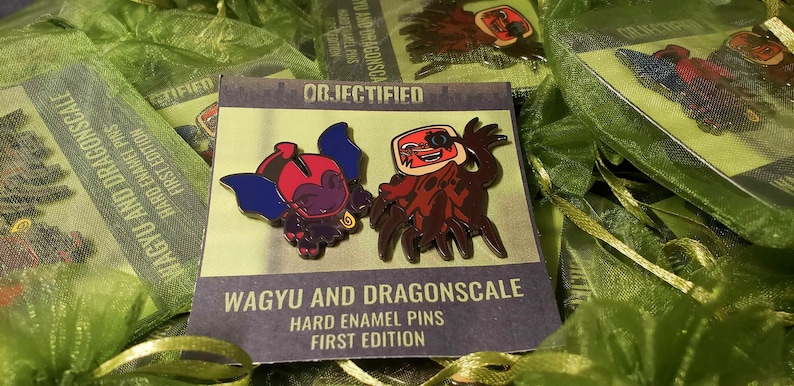 May include: Hard enamel pins featuring two cartoon characters, one with blue wings and the other with a red and brown design. The pins are on a card that reads "WAGYU AND DRAGONSCALE HARD ENAMEL PINS FIRST EDITION". The pins are packaged in green mesh bags.