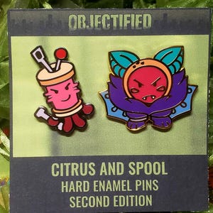 May include: Two hard enamel pins on a display card labeled OBJECTIFIED CITRUS AND SPOOL HARD ENAMEL PINS SECOND EDITION. One pin is a pink spool with needles, the other is a purple and orange citrus character with leaves. Both have gold metal outlines.