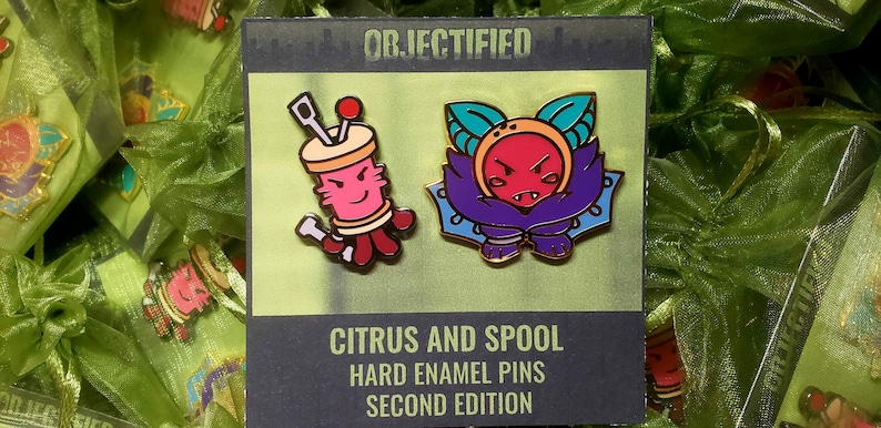 May include: Two hard enamel pins on a display card labeled OBJECTIFIED CITRUS AND SPOOL HARD ENAMEL PINS SECOND EDITION. One pin is a pink spool with needles, the other is a purple and orange citrus character with leaves. Both have gold metal outlines.