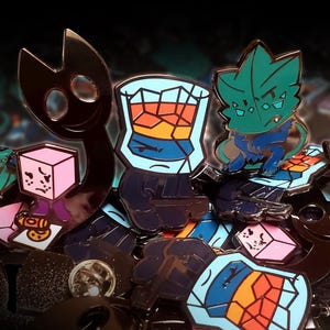 Puede incluir: Una colecci&oacute;n de pines de esmalte met&aacute;lico con varios dise&ntilde;os, incluyendo una criatura de silueta oscura, un personaje de cubo rosa, un vaso lleno de l&iacute;quido naranja y azul, y una criatura verde azulada con detalles en azul, con cierres.