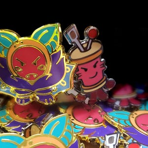 May include: A collection of metal enamel pins featuring two distinct designs. One pin depicts a stylized creature with a red face, purple body, and teal leaf-like ears. The second pin shows a pink cylindrical character with tools on top.