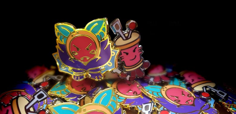 May include: A collection of metal enamel pins featuring two distinct designs. One pin depicts a stylized creature with a red face, purple body, and teal leaf-like ears. The second pin shows a pink cylindrical character with tools on top.