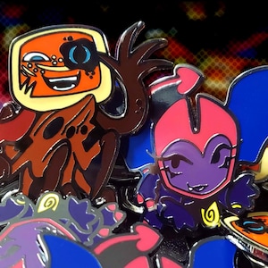 May include: A collection of enamel pins featuring cartoon characters. One pin has an orange face with a blue eye patch and a yellow border. Another pin has a purple character with a pink head and blue wings. Other pins are visible.