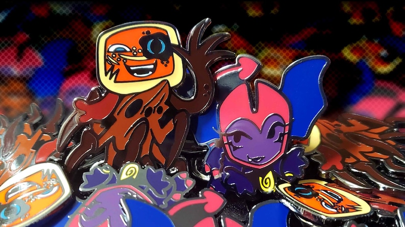 May include: A collection of enamel pins featuring cartoon characters. One pin has an orange face with a blue eye patch and a yellow border. Another pin has a purple character with a pink head and blue wings. Other pins are visible.