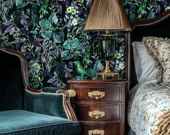 May include: A dark green velvet armchair with gold trim sits next to a wooden nightstand with three drawers and gold handles. A green bird figurine sits on the nightstand next to a gold table lamp with a brown lampshade. The wall behind the furniture is covered in a dark green and purple floral wallpaper.