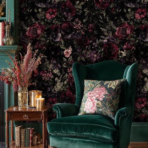 May include: A dark green velvet armchair with a floral patterned pillow sits in front of a floral wallpaper. A small wooden side table holds candles, books, and a vase of flowers. A patterned rug is on the floor.