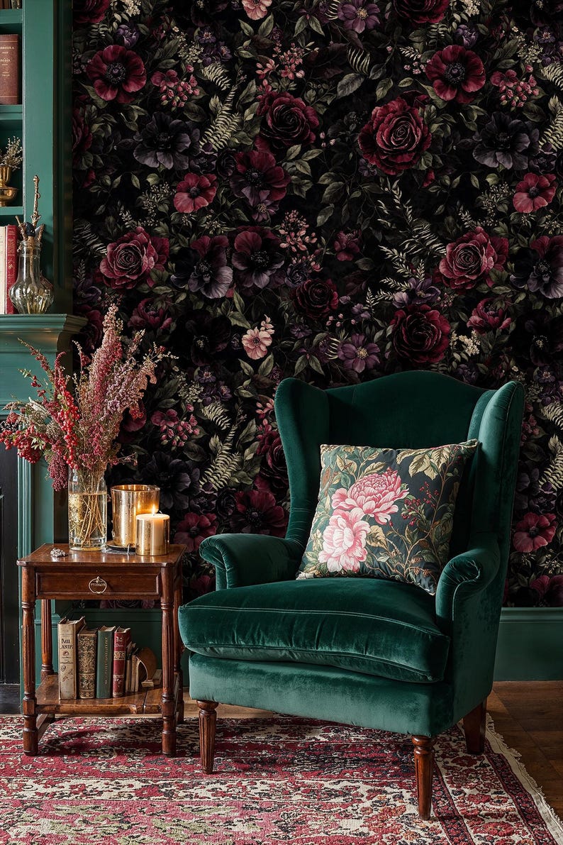 May include: A dark green velvet armchair with a floral patterned pillow sits in front of a floral wallpaper. A small wooden side table holds candles, books, and a vase of flowers. A patterned rug is on the floor.