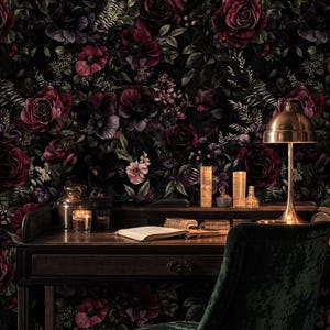 May include: A dark, moody interior scene featuring a wooden desk with a brass lamp, candles, and an open book. The background is a floral wallpaper with deep red and purple roses. A green velvet chair completes the scene.
