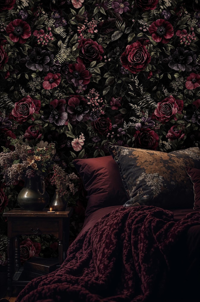 May include: A dark, moody bedroom scene featuring a floral wallpaper with deep red roses and other flowers. A bed with burgundy bedding, pillows, and a knitted throw is in the foreground. A vase of flowers and a candle sit on a wooden nightstand.