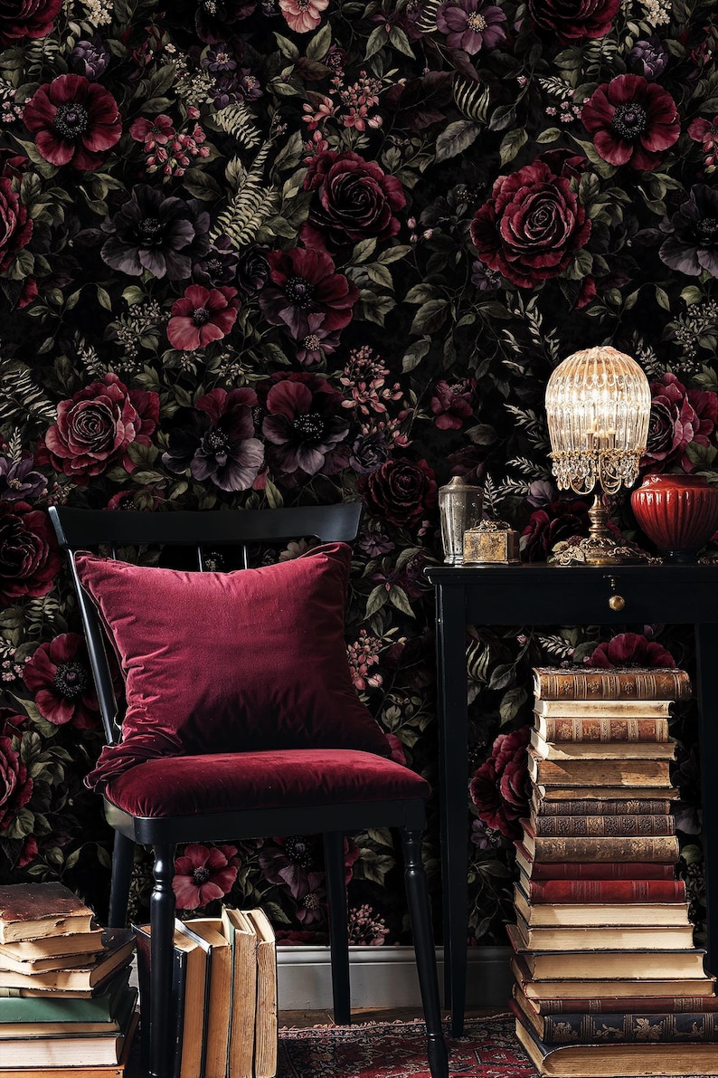 May include: A dark, floral wallpaper featuring burgundy and dark purple roses and other flowers. A black chair with a burgundy velvet cushion and seat is in front of the wallpaper. A small black table holds a lamp and books.