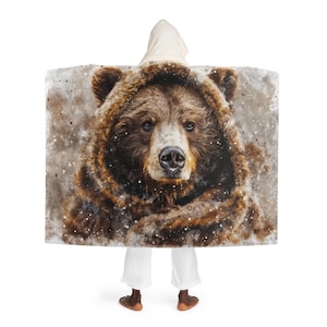 Puede incluir: Una manta con capucha con una pintura de acuarela de la cara de un oso pardo. El oso tiene un hocico marrón claro y ojos oscuros. La manta es marrón y beige con una capucha blanca.