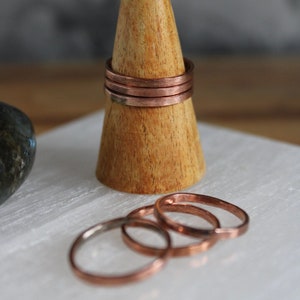 May include: Copper rings on a wooden cone stand. Several rings are stacked on the stand, with others scattered nearby. The rings have a simple design and a warm, reddish-brown colour. The rings are a minimalist style.