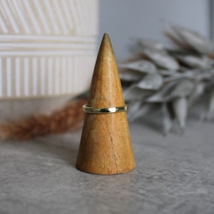 Könnte beinhalten: Ein goldener Ring, der auf einem Holzringhalter präsentiert wird. Der Ring hat ein strukturiertes Band. Der Ringhalter ist ein hellbraunes, kegelförmiges Objekt. Der Hintergrund ist ein neutrales Grau.