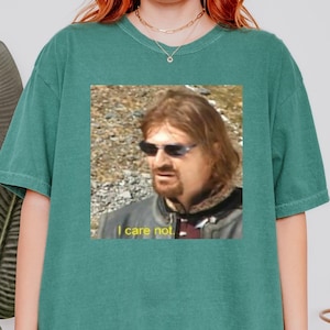 Op de afbeelding: Een lichtgroen T-shirt met een afbeelding van een man met een zonnebril en een zwart jack. De tekst "I care not." is onder de afbeelding gedrukt.