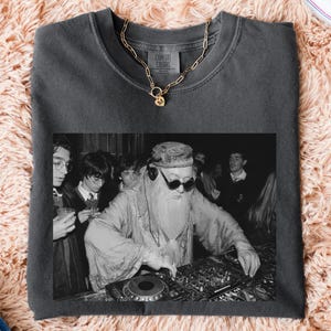 May include: A charcoal gray crewneck sweatshirt featuring a black and white graphic print of a bearded figure wearing sunglasses and a hat while operating DJ equipment, accented by a gold-toned chain necklace resting on the collar.