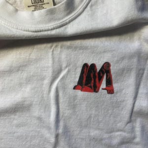 Puede incluir: Camiseta blanca con un gráfico rojo y negro de una persona sentada con las piernas cruzadas. La camiseta tiene una etiqueta que dice "Comfort Colors" y "Heavyweight Garment Dye".