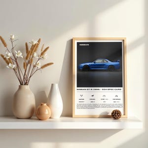 Puede incluir: Impresión enmarcada de un coche Nissan GT-R (1999) azul, con especificaciones y el logotipo de Nissan. El marco es de madera clara, expuesto en una estantería blanca con jarrones. Una elegante pieza de arte automovilístico.