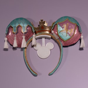 Könnte beinhalten: Ein Paar rosa und türkisfarbene Mäuseohren mit einer goldenen Krone und einem Schild mit einem Karussellpferd-Design. Die Ohren sind mit Pailletten und Quasten verziert.