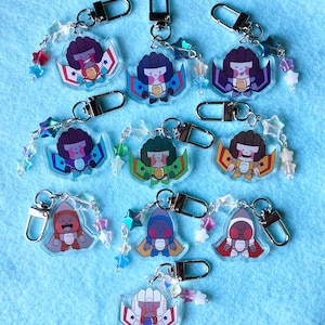 以下が含まれることがあります： ジェット機のような衣装を着た漫画のキャラクターが特徴の、カラフルなアクリルキーホルダーのコレクション。各キーホルダーにはシルバーの留め金が付いており、さまざまな色の星型のビーズのストリングで飾られています。キャラクターは、さまざまな色の翼と衣装を着ています。
