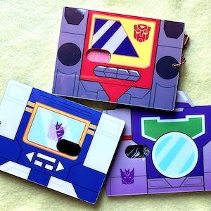May include: Three colorful, rectangular Transformers-themed cardholders. The designs feature the characters' iconic looks, with details like buttons, screens, and the Autobot or Decepticon symbols. Each cardholder has a small metal chain.