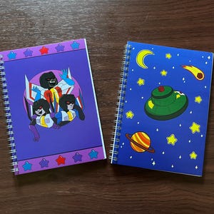 May include: Two spiral bound notebooks with different designs. The first notebook has a purple cover with a graphic of three cartoon characters in a spaceship. The second notebook has a blue cover with a graphic of a spaceship, stars, and planets.