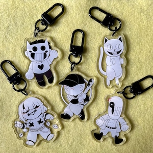 Puede incluir: Cinco llaveros de personajes de dibujos animados en blanco y negro con llaveros de metal negro. Los personajes incluyen un gato, un jugador de béisbol, una persona con un corazón en la camisa, una persona con una calavera en la camisa y una persona con una calavera en la cabeza.