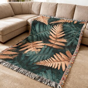 May include: A woven throw blanket with a fern leaf pattern. The blanket has a dark green backdrop with large, tan-coloured fern leaves. The edges are finished with a fringe in black, white, and colourful threads. The blanket is draped over a beige sofa.