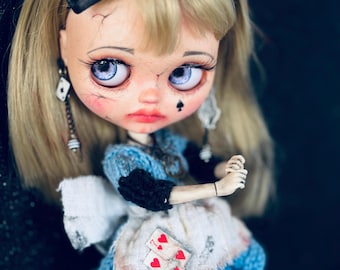 May include: A doll with blonde hair, a black hair bow, and large blue eyes. The doll wears a blue knitted dress with a white apron featuring two red heart playing cards. It has pale skin with painted cracks, a black spade mark on the cheek, and dangling playing card earrings.