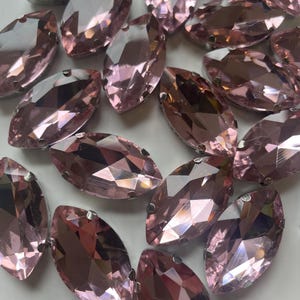 May include: A collection of marquise-shaped, faceted rhinestones in a soft pink hue. Each stone has multiple facets that reflect light, creating a sparkling effect. The rhinestones are arranged closely together, showcasing their individual shapes and colors.