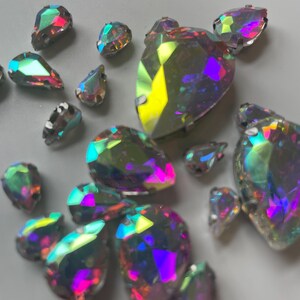 May include: A collection of iridescent rhinestones in various shapes and sizes. The rhinestones reflect a spectrum of colors, including pink, purple, yellow, and green. The shapes include teardrops, hearts, and other geometric forms.
