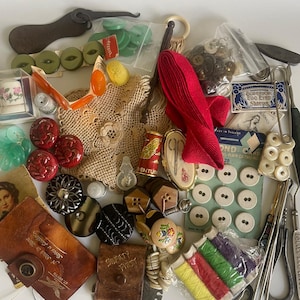 Puede incluir: Una colección de nociones de costura vintage, que incluye botones, dedales, carretes de hilo y una cinta roja. Se ven varios estilos de botones, junto con un pequeño estuche de cuero y un abridor de costuras de metal. Los artículos están dispuestos sobre una superficie blanca.