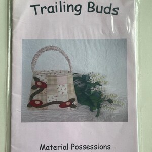 May include: A pink and white fabric basket with a brown handle and red flower accents. The basket is labeled "Trailing Buds" and "Material Possessions Patchwork & Quilting www.materialpossessions.com.au".