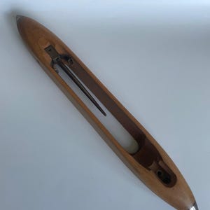 Op de afbeelding: Een houten weefspoel, ongeveer 30 cm lang, met een puntig uiteinde en een metalen punt. Het heeft een holle binnenkant en een metalen pin. Het hout is lichtbruin.