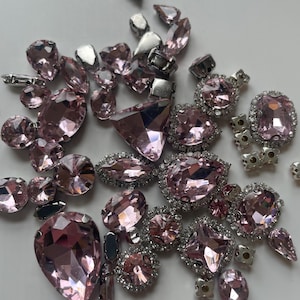 May include: A collection of pink rhinestones in various shapes and sizes, including teardrops, ovals, and faceted gems. Some rhinestones are set in silver-colored metal settings, and others are loose. The gems have a sparkling, reflective quality.