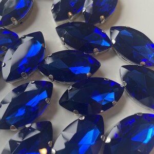 May include: A collection of marquise-shaped, faceted, deep blue glass rhinestones. Each gem is set in a silver-colored metal setting. The gems are arranged on a white surface, reflecting light and creating a sparkling effect. These are likely used for embellishments.