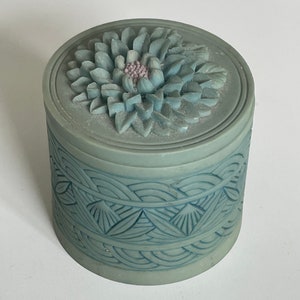 May include: A round, light blue trinket box with a floral design on the lid. The lid features a detailed, three-dimensional flower with a pink center. The box's body is adorned with a repeating leaf and wave pattern.